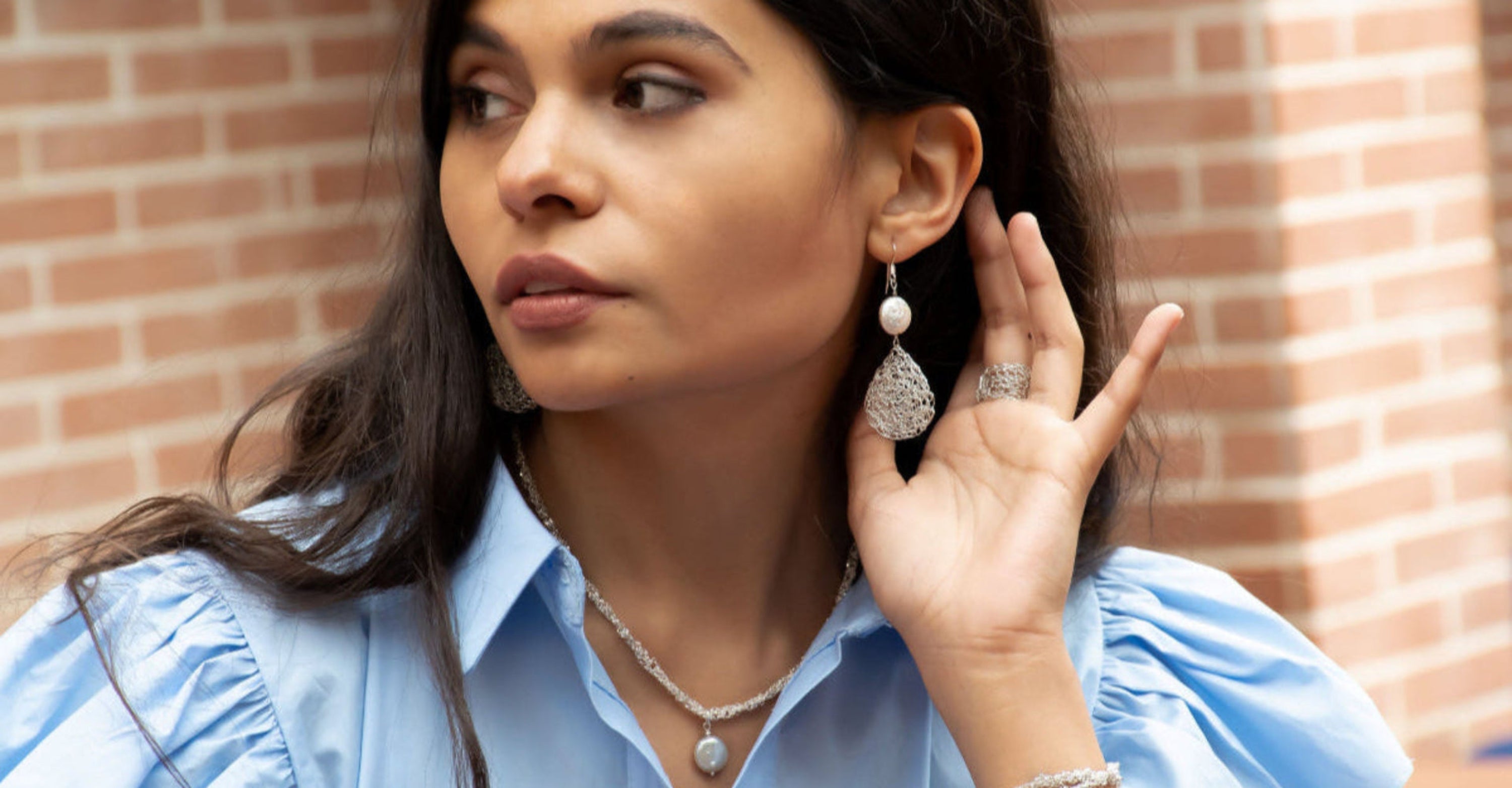 Woman wearing pearl drop earrings and a pearl necklace, standing against a brick wall.