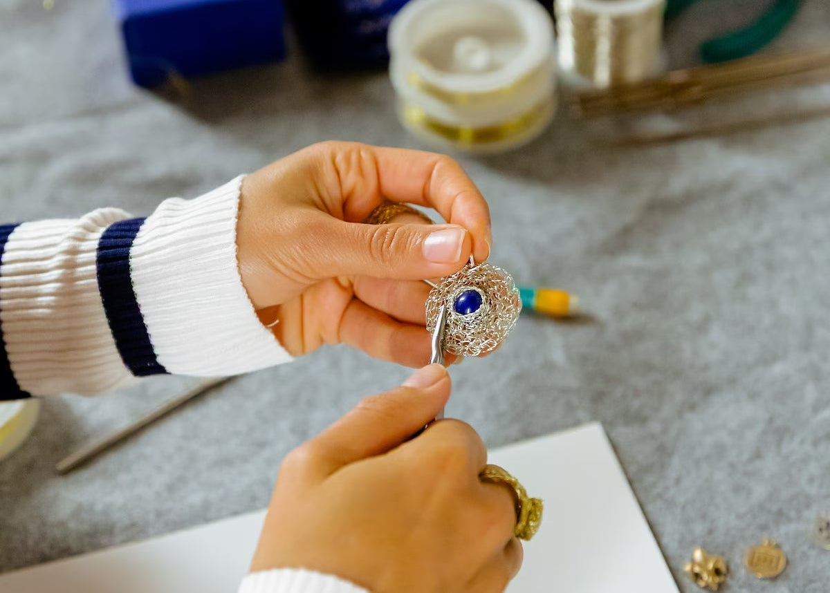 Person holding a small wire knitted charm with a jewelry-making setup in the background.
