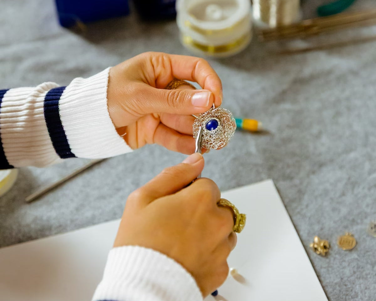 Maria Glezelli holding a silver piece with a blue lapis on a textured surface with tools and materials.